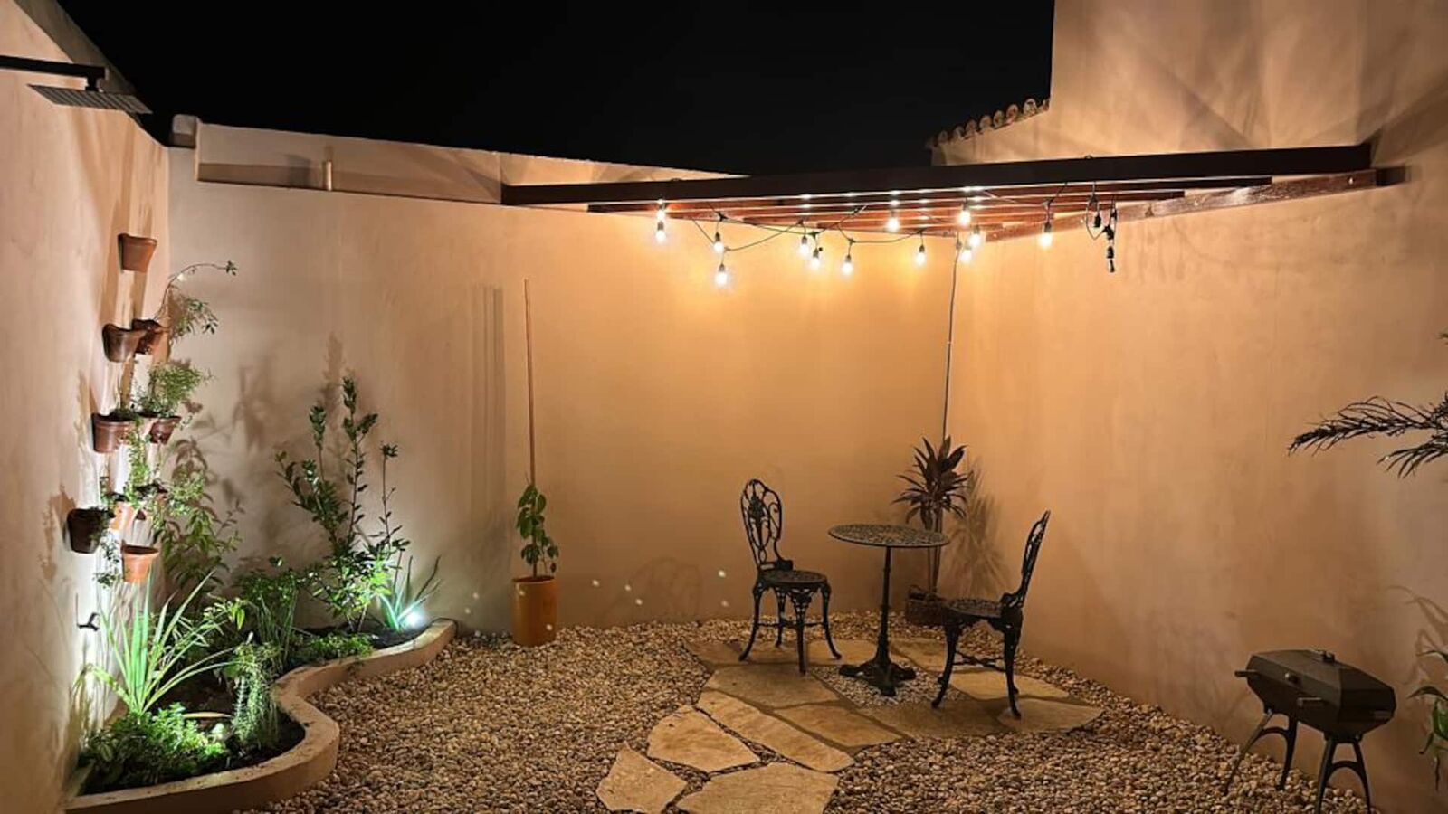 Pátio da Casa Boutique à noite, com luzes pendentes, mesa de ferro e espreguiçadeira entre as plantas